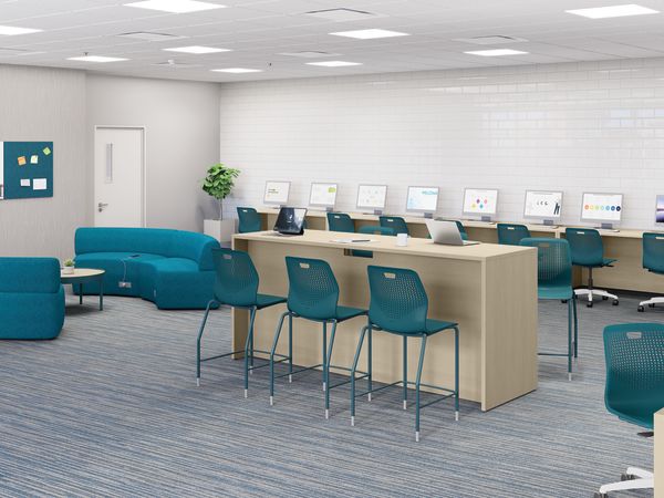 Modern computer lab with long worktables, desktop monitors, laptops, teal task chairs, soft seating area, white brick accent wall, carpeted flooring, and bright overhead lighting.