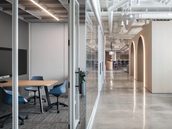 Glass-enclosed meeting room with round wood table, rolling task chairs, wall-mounted display, and adjacent lounge seating with additional tables and chairs.