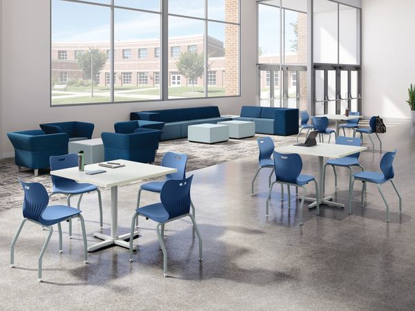 Bright student commons area with collaborative tables, blue classroom chairs, lounge seating, floor-to-ceiling windows, polished concrete flooring, and open gathering space in a modern educational environment.