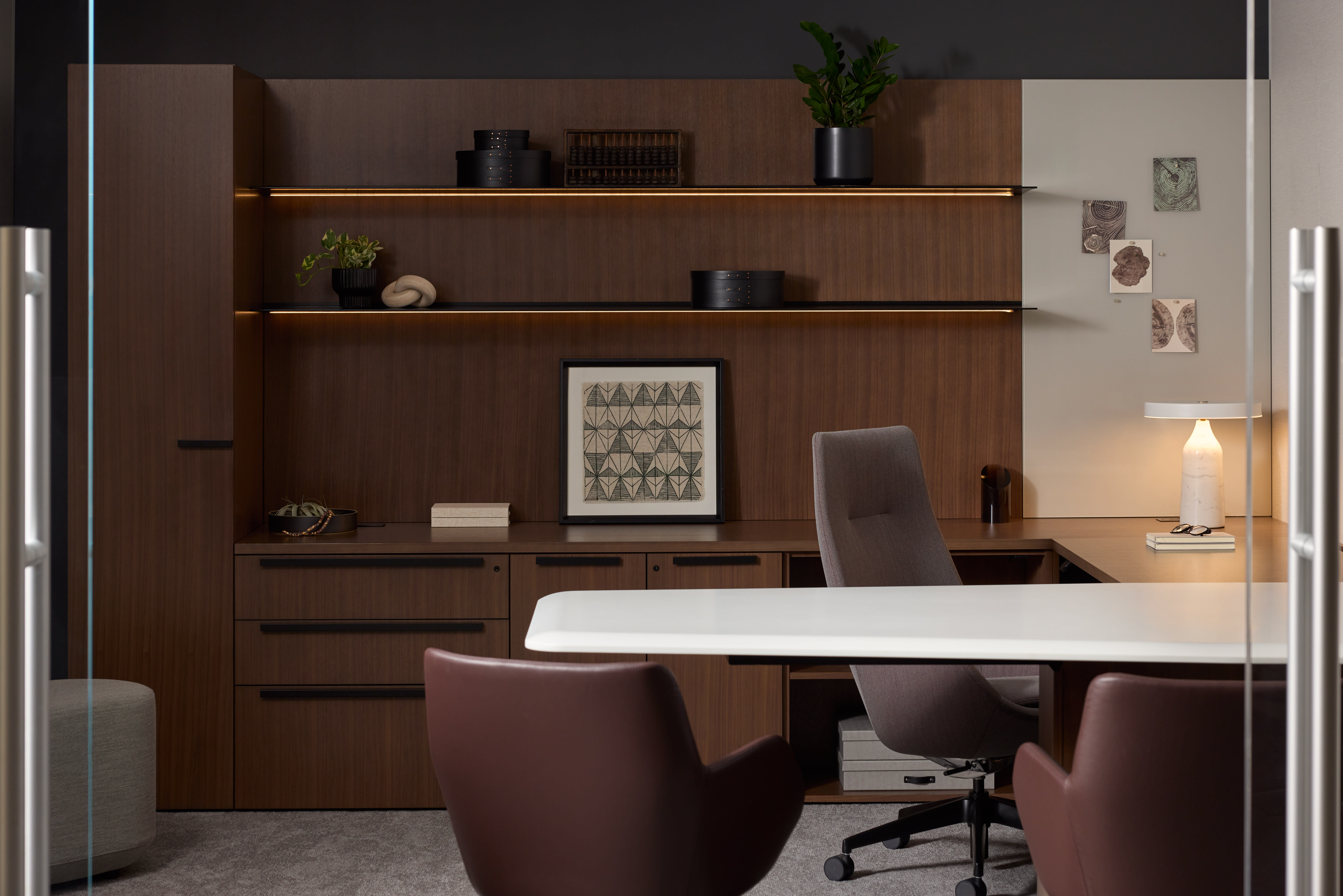 Private office with wood storage and display shelves on back wall next to an L-shaped desk with a high back task chair and two guest chairs pulled up.