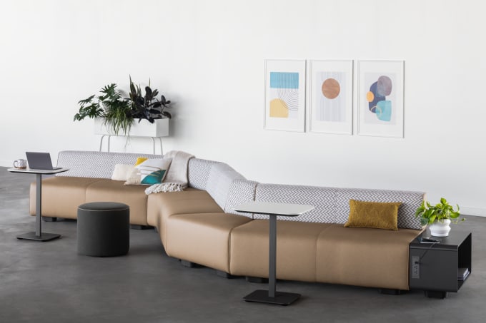 Tan modular lounge seating in serpentine formation with herringbone back material, two laptop tables, a grey pouf, and white planter in the background.