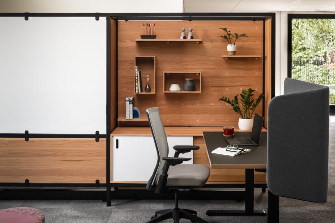 Modern height adjustable workstation with grey fabric screen, dark grey task chair, with wood storage, shelves, and markerboard adjacent to it.