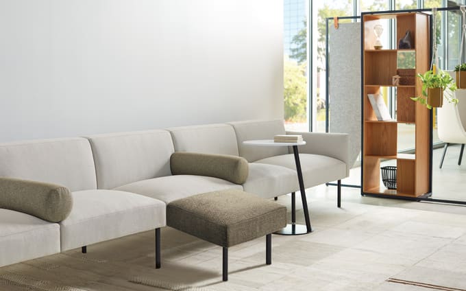 Low-profile lounge system in light upholstery with tan bolsters and ottoman with bookcase space division to the right of the sofa.