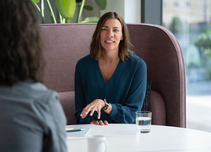 Two people seated at a white round table in a modern office lounge with maroon upholstered booth seating and large windows providing natural light.