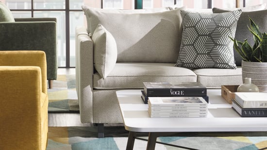 A cozy and modern living room with a beige sofa, patterned throw pillows, a yellow armchair, and a glass coffee table on a colorful area rug, set against a backdrop of large windows overlooking a city skyline.