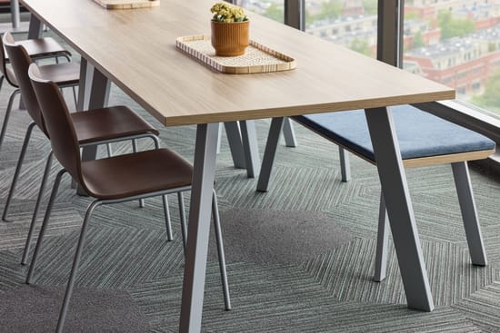 A modern, minimalist dining table with a wooden top and metal legs sits in a room with a patterned carpet and large windows overlooking an outdoor area.