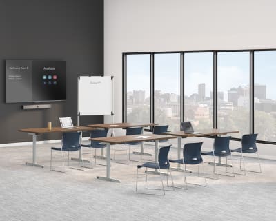 A modern training room with long tables, blue chairs, a wall-mounted display screen, a whiteboard, and large windows overlooking a city skyline.