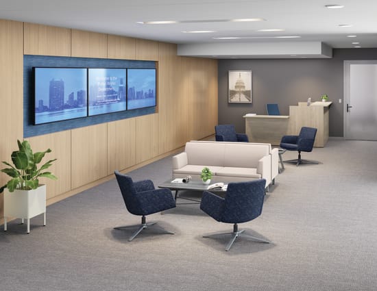 Modern office lounge with white sofa, blue lounge chairs, wall-mounted digital display, wood paneling, and reception desk.