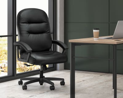 A black leather office chair sits in front of a large window, with a wooden desk and laptop visible in the background.