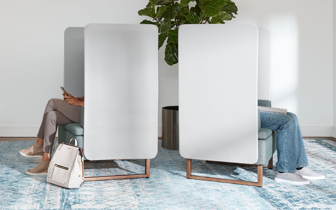 The image depicts a minimalist, modern interior design with two white, freestanding panels or screens placed in the foreground, surrounded by various household items such as a potted plant, a bag, and some clothing on the floor.