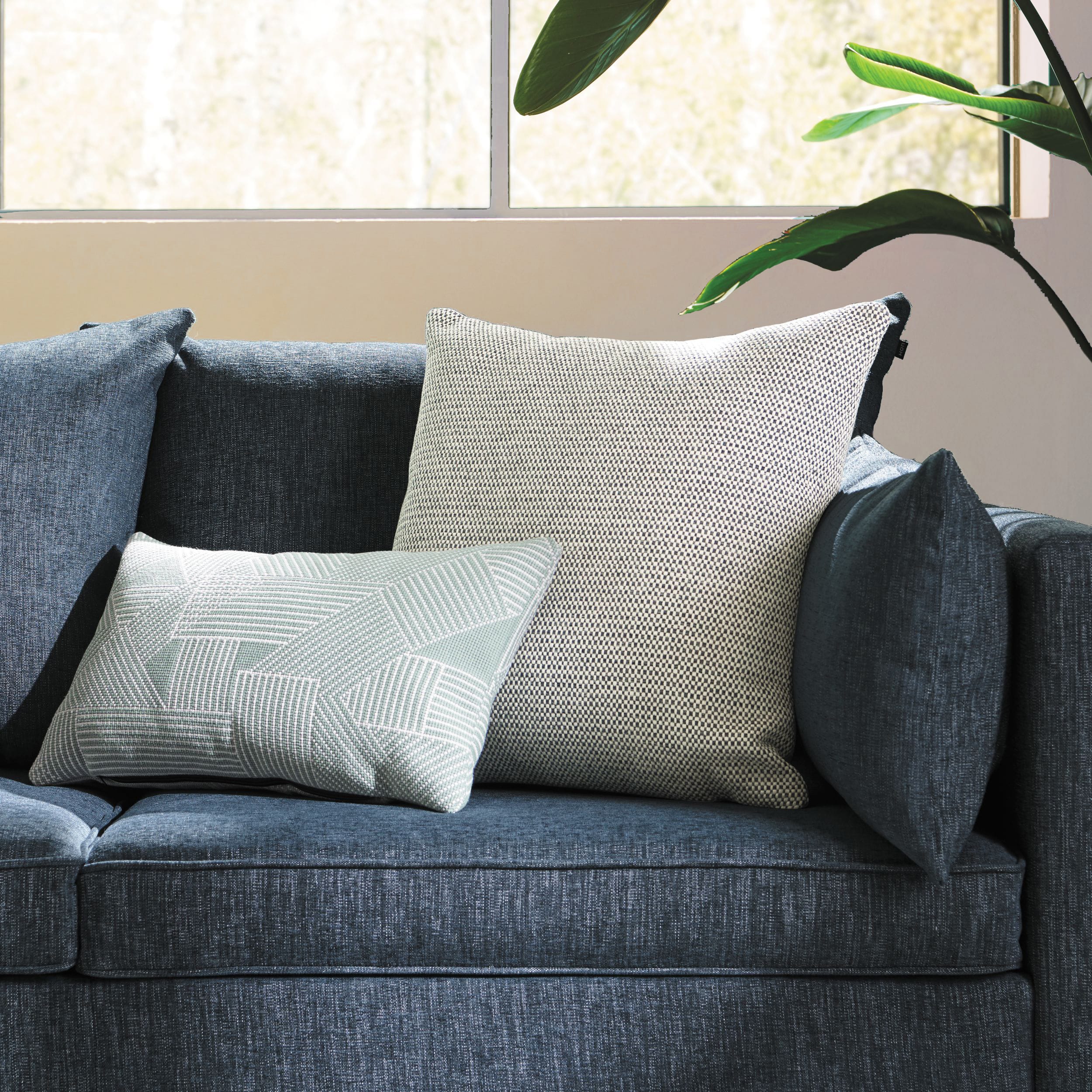 A cozy living room with a gray sofa, patterned throw pillows, and a large window overlooking lush greenery.
