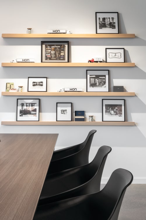 Copilot said: Conference space with wood meeting table, molded guest chairs, glass partition wall, and floating wood shelves displaying framed prints and accessories.