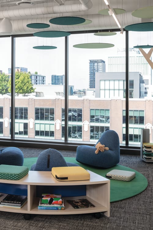 Flexible lounge area with upholstered seating, ottomans, low tables, open shelving, and acoustic ceiling panels near large windows.