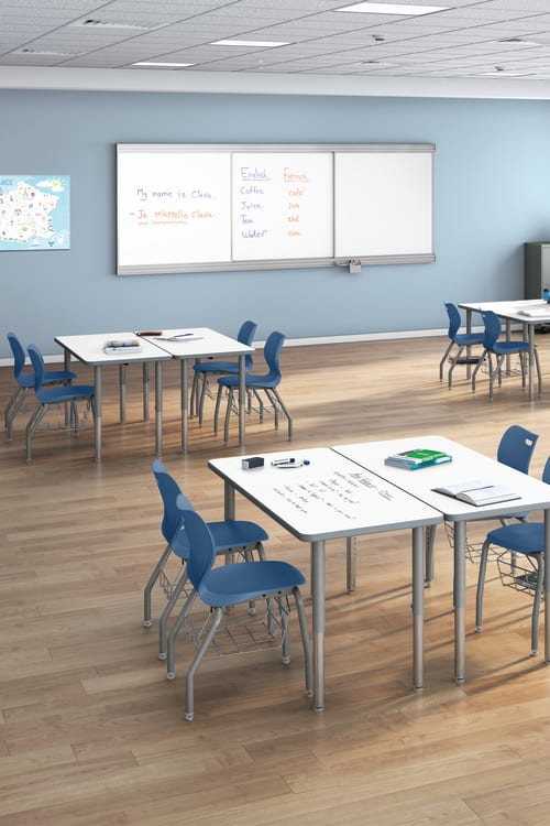 Modern classroom with grouped tables, blue chairs, large windows, educational posters, map of France, and whiteboard displaying English and French vocabulary.