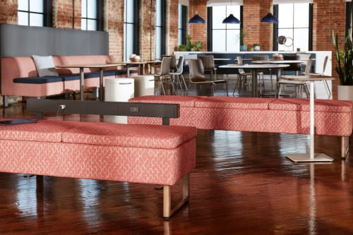 A modern and stylish office space with brick walls, large windows, and a mix of comfortable seating options, including plush pink sofas and wooden chairs, along with a long wooden table in the background.