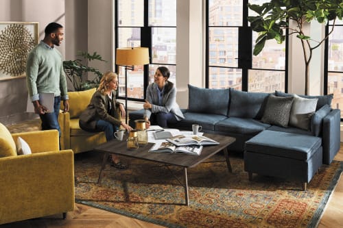 The image depicts a cozy living room with a group of people sitting on a blue sofa, surrounded by various decorative elements such as plants, artwork, and a patterned rug.