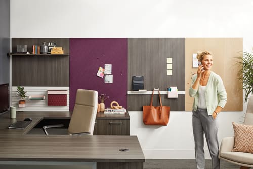 A woman stands in a modern, well-furnished home office, surrounded by shelves, a desk, and various decor items.
