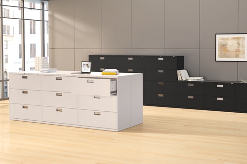 The image depicts a modern office space with a white filing cabinet and a black storage unit in the foreground, set against a backdrop of large windows and framed artwork on the walls.