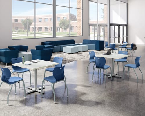 Bright student commons area with collaborative tables, blue classroom chairs, lounge seating, floor-to-ceiling windows, polished concrete flooring, and open gathering space in a modern educational environment.