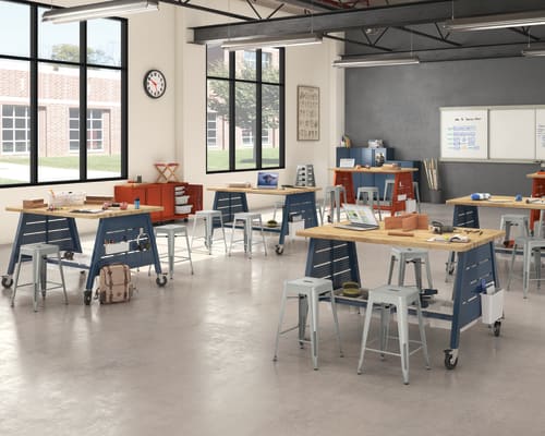 A modern, open-concept workspace with various workstations, desks, and chairs arranged throughout the room. The space features large windows, exposed ceiling beams, and a whiteboard on the wall, creating a bright and collaborative environment.