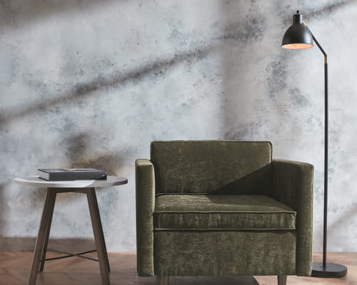 A cozy living room setting with a green upholstered armchair, a round wooden side table, and a floor lamp against a textured marble-like wall backdrop.