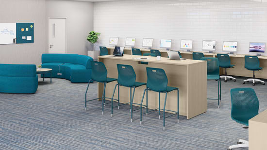 Modern computer lab with long worktables, desktop monitors, laptops, teal task chairs, soft seating area, white brick accent wall, carpeted flooring, and bright overhead lighting.