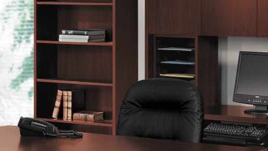 A modern and minimalist home office setup with a large wooden desk, bookshelves, and black office chairs arranged in a spacious room with a large window providing natural lighting.