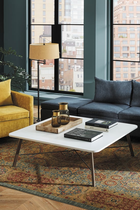 A cozy and stylish living room with a gray sectional sofa, yellow armchairs, a white coffee table, and a patterned area rug, set against a backdrop of large windows and lush greenery.