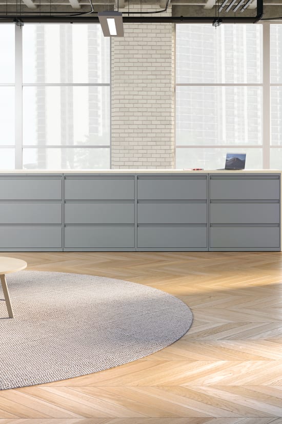 A modern, minimalist office space with gray storage units, yellow chairs, and a round rug on a hardwood floor, surrounded by large windows letting in natural light.