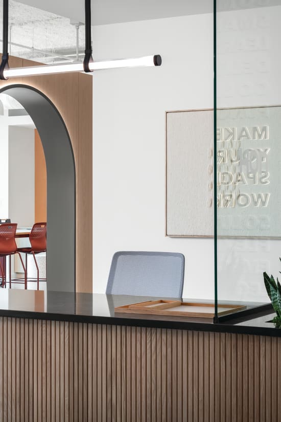 Reception counter with wood slat detailing and solid surface top, paired with task seating, integrated planter, pendant lighting, glass partition, and arched wall opening leading to collaborative seating area.