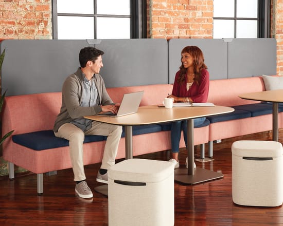 Two people, a man and a woman, are seated at a table in a modern office-like setting with brick walls and large windows in the background.