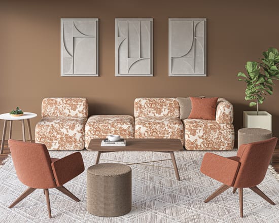 Contemporary lounge seating area with patterned sofas, upholstered lounge chairs, a low wood coffee table, and a textured area rug, styled against a warm brown accent wall with framed abstract wall art and a potted plant.
