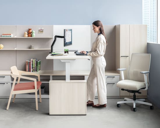 The image depicts a modern, minimalist home office setup with a woman standing at a standing desk, surrounded by shelves, chairs, and other office equipment.