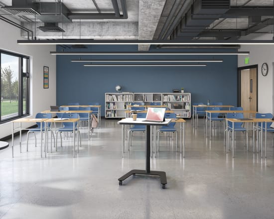 Modern classroom with flexible desks and chairs, large windows, blue accent wall, ceiling lighting, bookshelves, and teacher workstation with laptop and coffee cup.