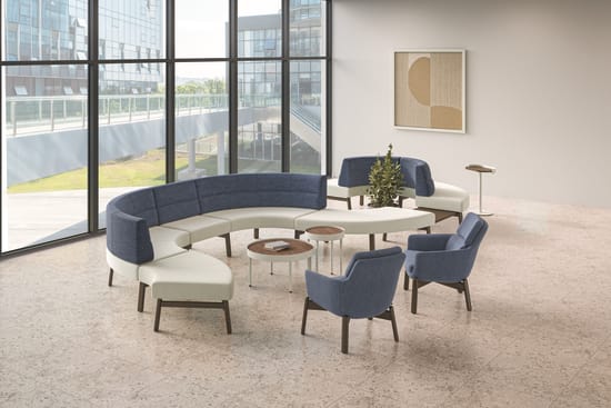 Modern lounge area with curved blue and white modular seating, blue lounge chairs, round wood accent tables, and floor-to-ceiling windows in an open collaborative workspace.