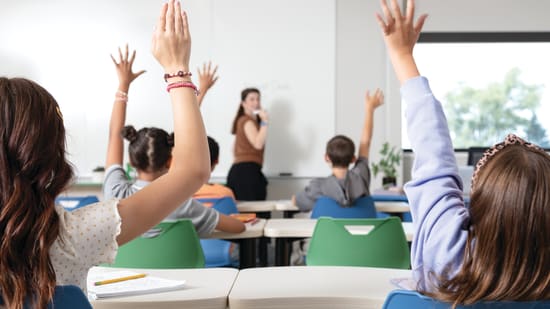 Students raising a hand during a classroom lesson with collaborative tables, green seating, and an instructor at the front of the room.