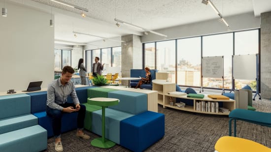Modular soft seating blocks with upholstered surfaces, paired with a small round pedestal table and low open shelving units for flexible workspace use.