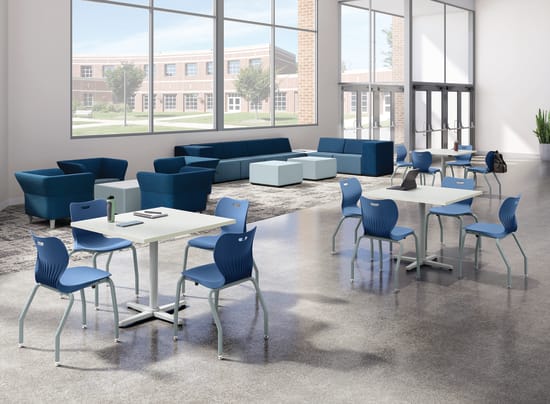 Bright student commons area with collaborative tables, blue classroom chairs, lounge seating, floor-to-ceiling windows, polished concrete flooring, and open gathering space in a modern educational environment.