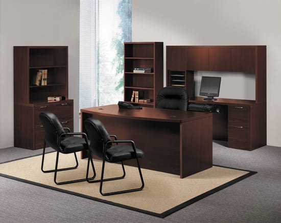 A modern and minimalist home office setup with a large wooden desk, bookshelves, and black office chairs arranged in a spacious room with a large window providing natural lighting.