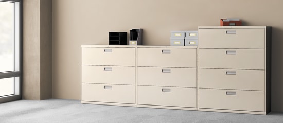 The image depicts a set of white filing cabinets in a minimalist office setting, with various office supplies and equipment visible in the background.
