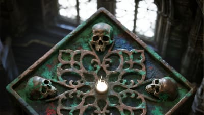 An ornate box with a textured green patina, decorated with metal skulls and intricate patterns, is illuminated by soft light.