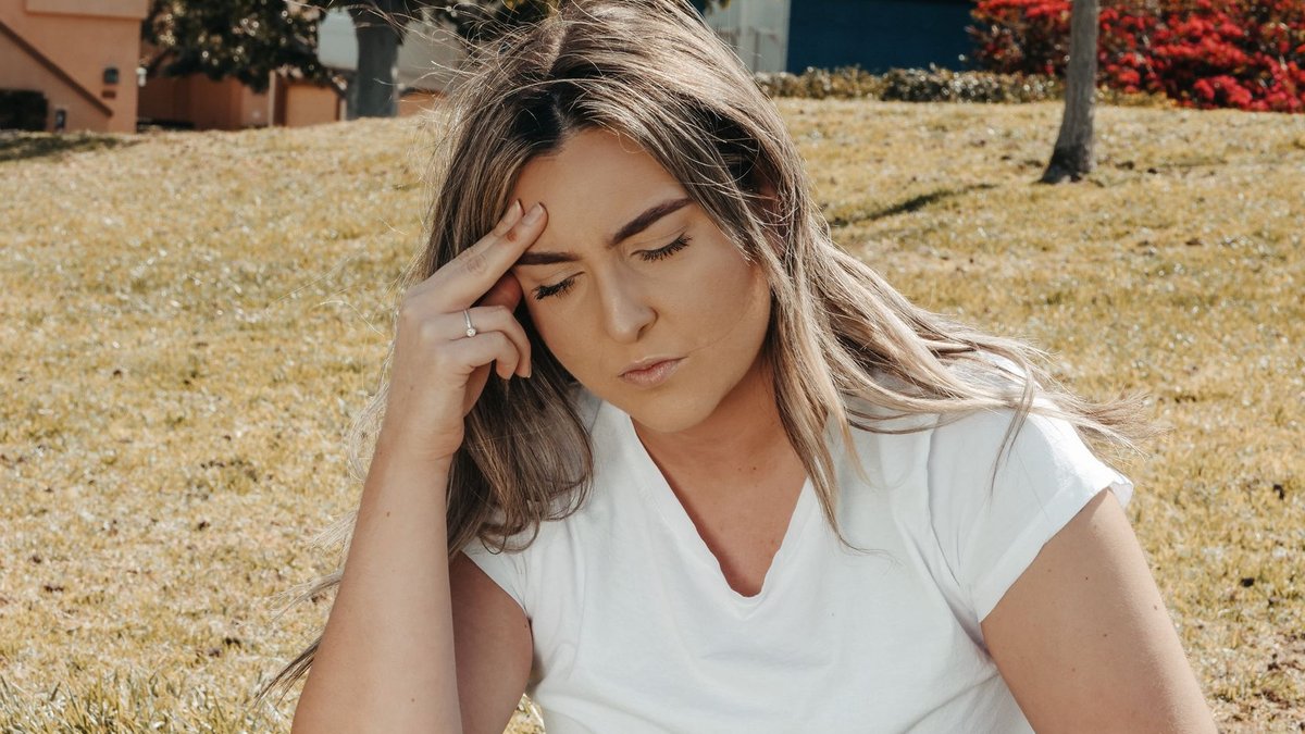 Neurologists Discuss Cluster Headache Treatments Caucasian woman sitting on grass outdoors with a headache, eyes closed, hand on head.