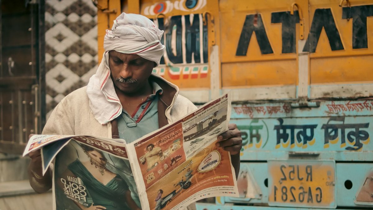 Man with mustache reads a newspaper next to a Tata dump truck on an urban street.