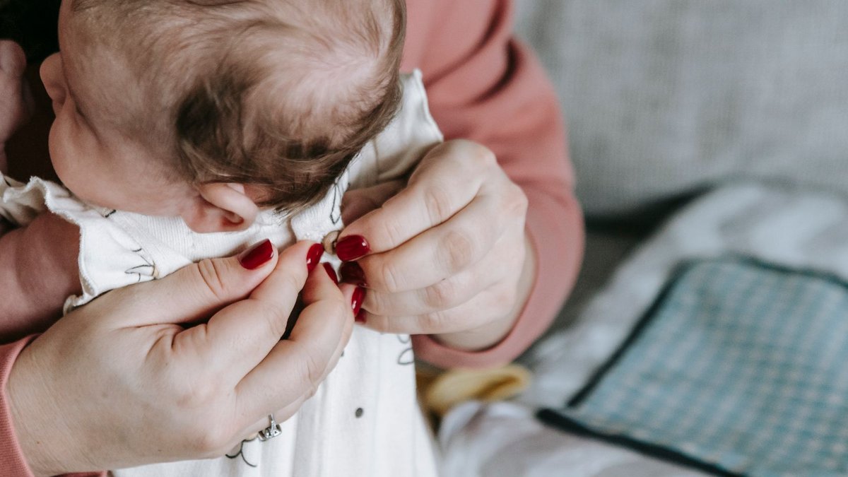 Close-up of a mother gently dressing her newborn baby indoors, highlighting tender caregiving moments.