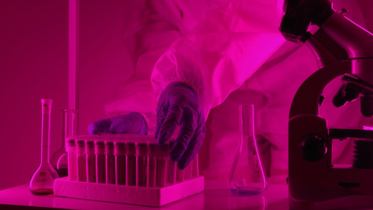 Sanofi invests $1 billion to boost Regeneron partnership and next-gen Scientist in a lab coat handling test tubes under pink lighting, using a microscope.