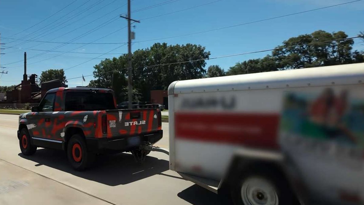 Camouflaged Slate electric pickup towing a U-Haul cargo trailer on a public road during testing.