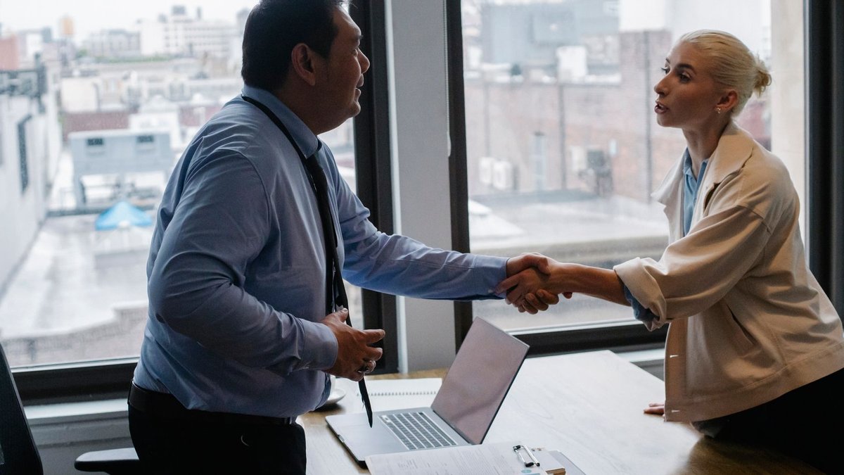 Young female candidate shaking hands with employer before job interview in light office with urban landscape through windows.