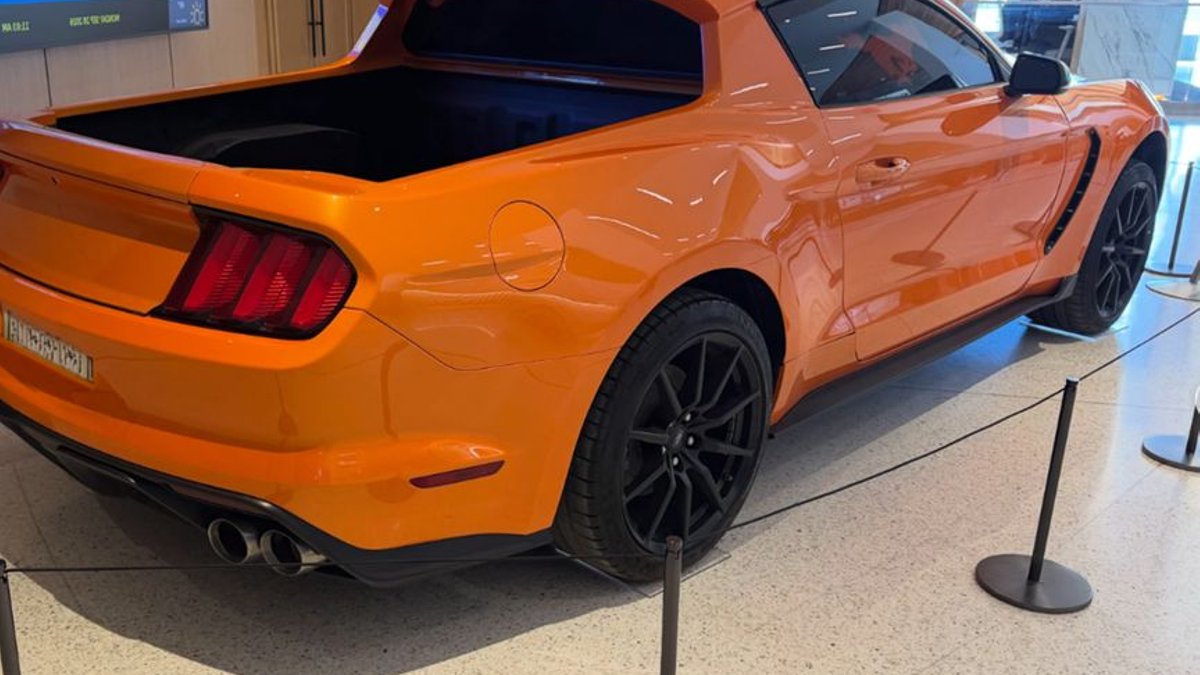 Ford Unveils One-Off Mustang Ute Concept, No Production Plans An orange Ford Mustang converted into a ute with a tray at the back, displayed behind rope barriers in a building foyer, seen