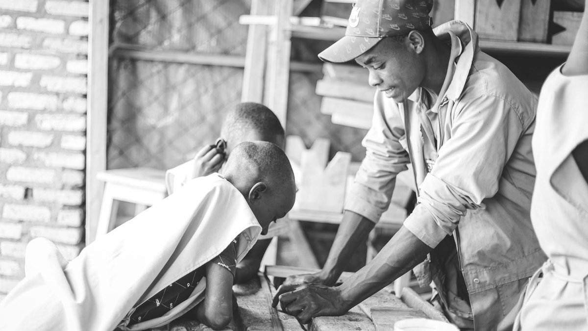 A mentor guides young boys in a carpentry workshop, fostering skill and curiosity in woodworking.