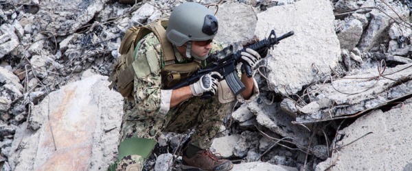 A special forces officer hiding amongst rocks, carrying a rifle.